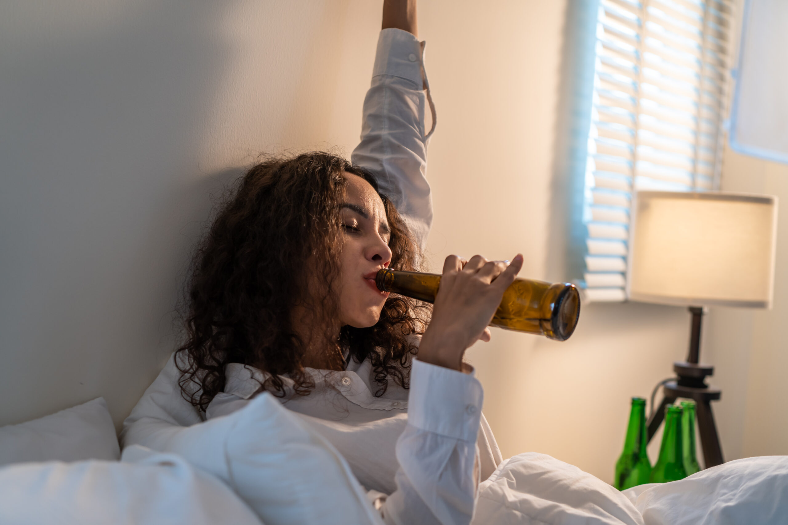 Drunk Latino woman hold beer bottle, feel hangover and dancing on bed. Young beautiful happy female stting in dark night in bedroom, feel alone and celebrate life problem with alcohol party at house.