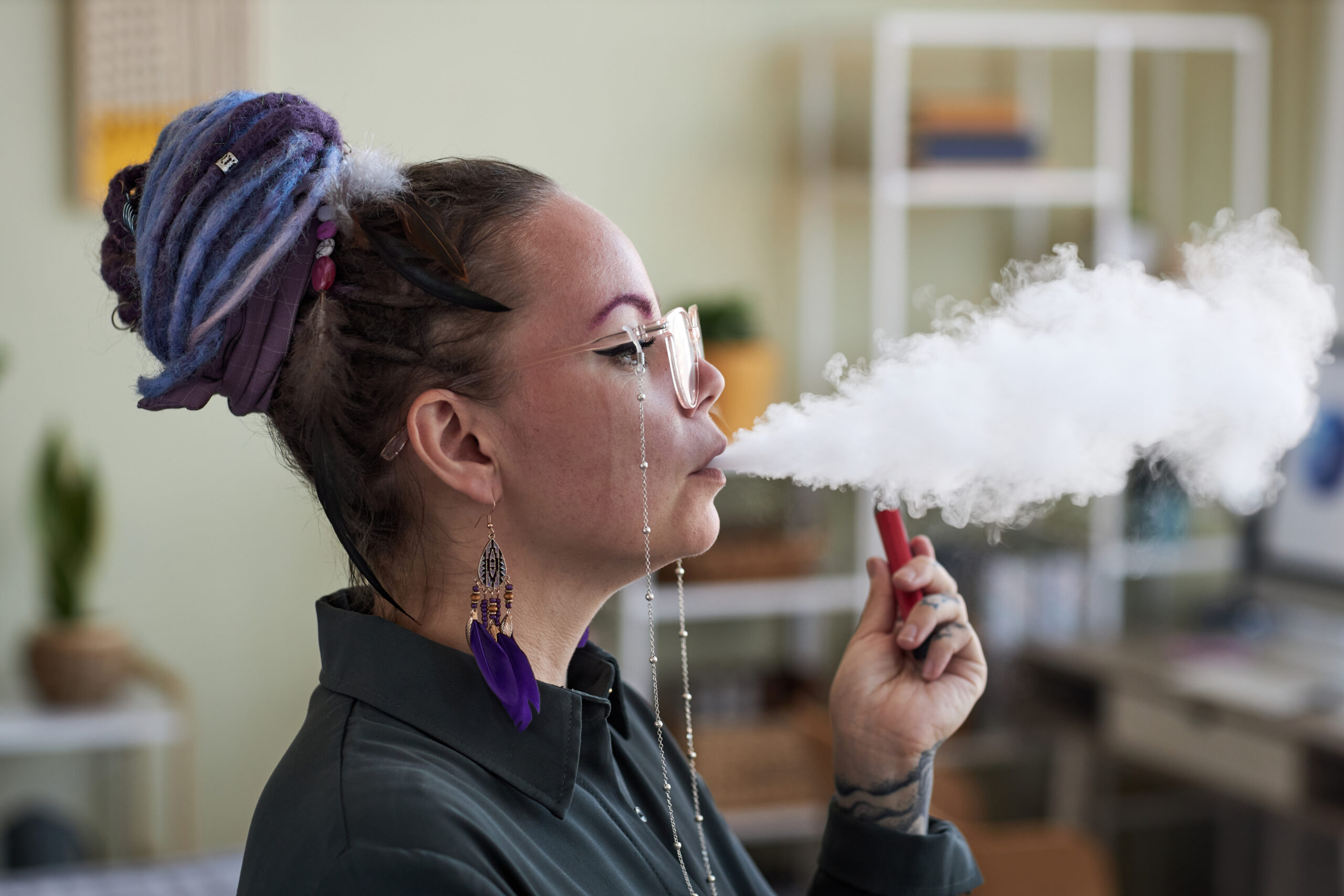 Side view of young woman in eyeglasses blowing white cloud of thick smoke out of her mouth while smoking in front of camera