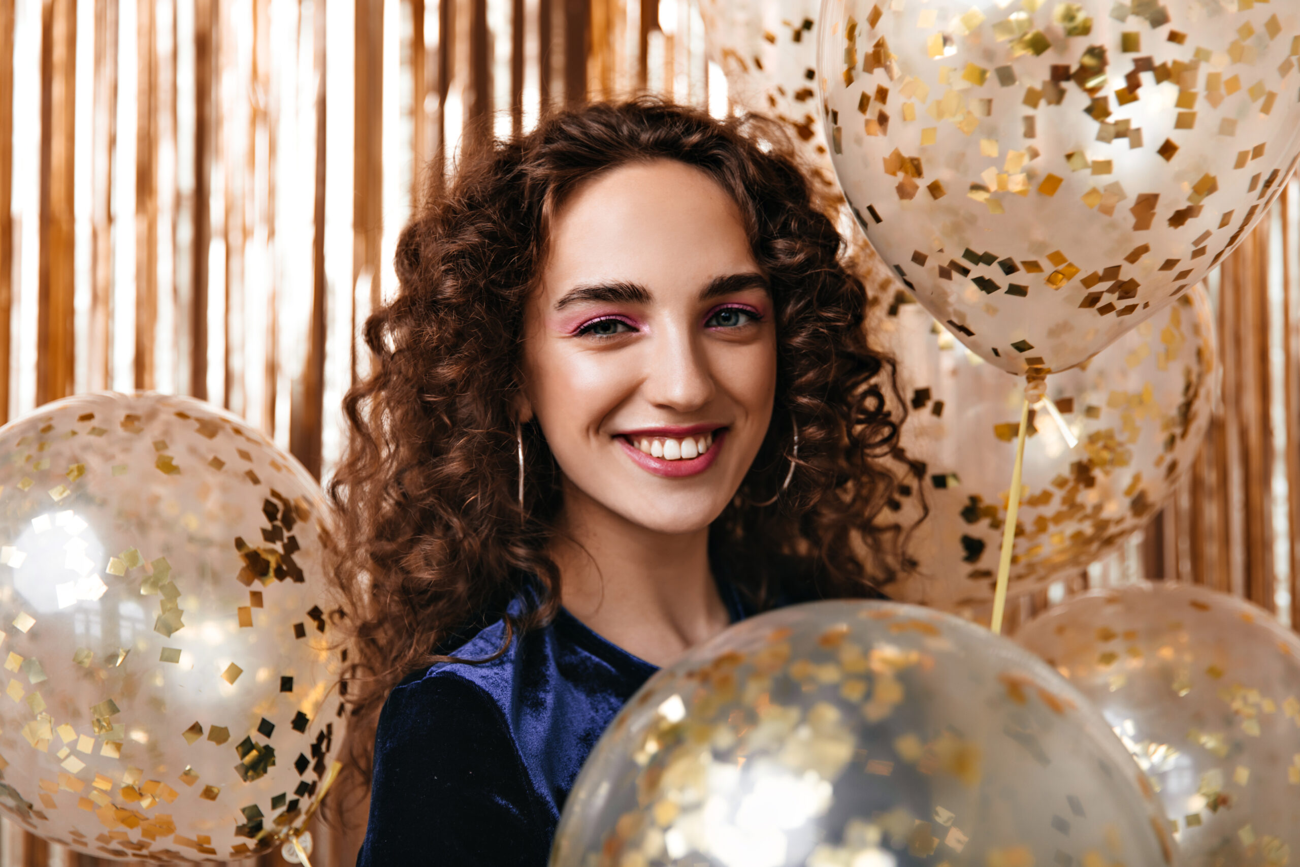 Beautiful girl with pink eyeshadows winks on gold background with balloons
