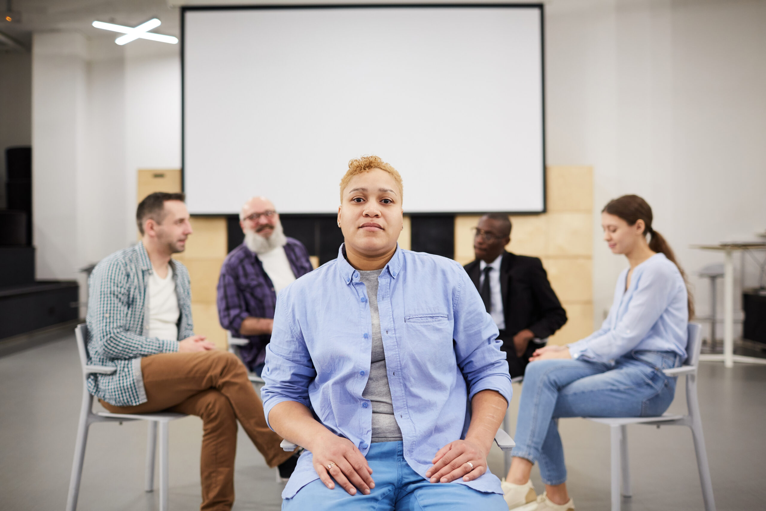 Portrait of masculine woman looking at camera while sitting in chair in front of therapy group, copy space