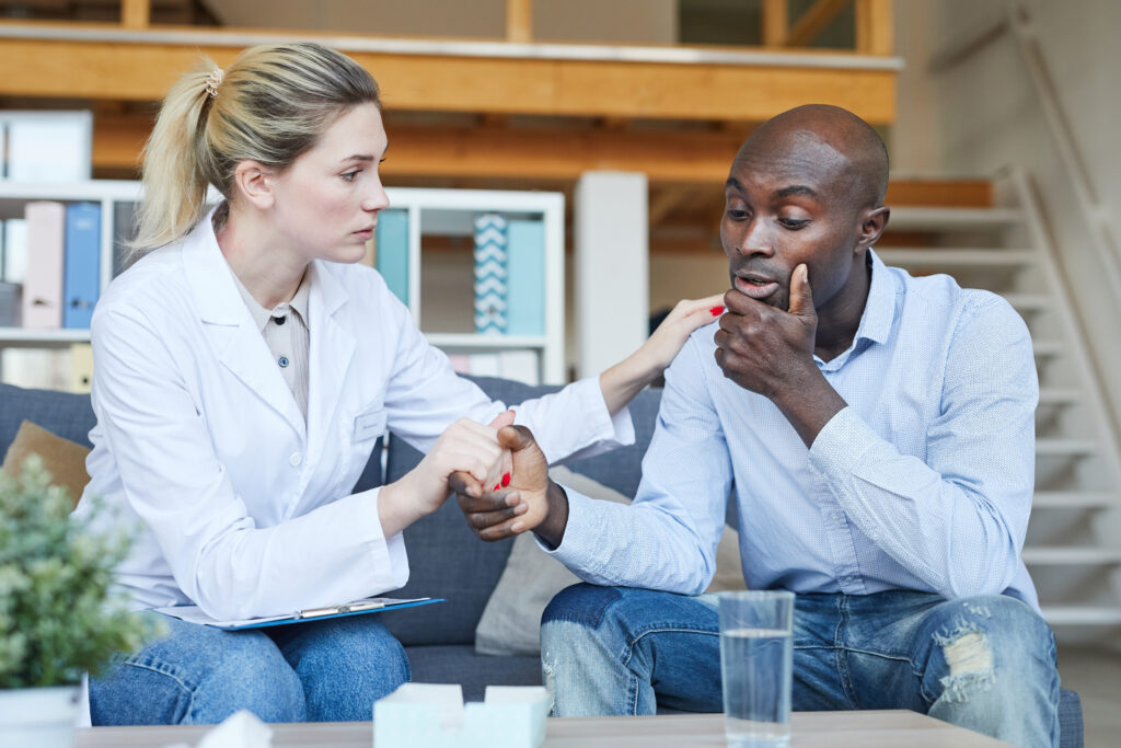 Understanding female therapist with badge on white coat sitting on sofa and supporting Afro-American patient at psychology session