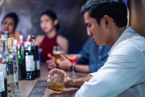 Asian depression man feeling heart broken and drinking beer in a bar. Attractive male sitting on counter bar, holding a bottle of alcohol feeling loneliness, drunk and hangover alone at night club.