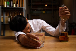 The drunk man wearing white shirt sleeping on the table holding the glass and bottle of whiskey that put on the wooden table in restaurant. Selective focus shot on the glass. Drunk man concept.