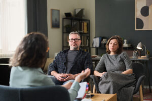 Couple sitting in office discussing relationship issues with therapist holding clipboard. Room has bookshelf, lamp, abstract painting, and large window providing natural light