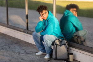 A thoughtful young brazilian guy sits by a glass pane, reflecting on life as their image casts back at them, exuding a sense of introspection and solitude