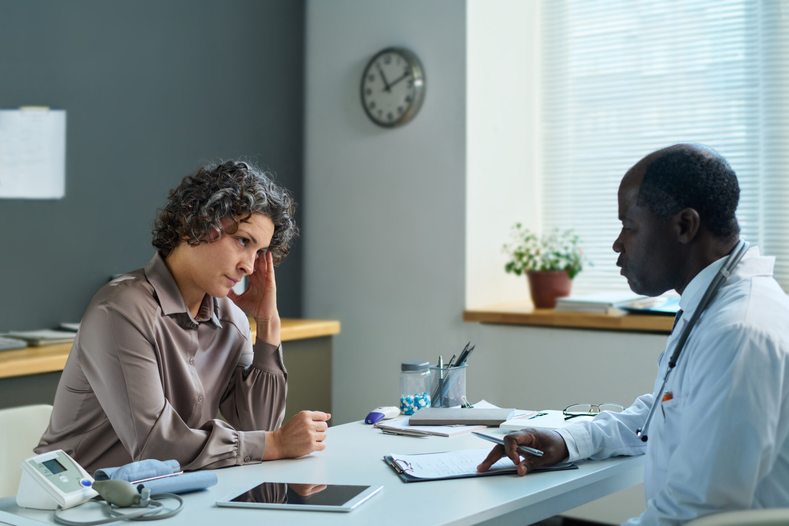 Serious mature woman touching head while sitting in front of African American male general practitioner and listening to him