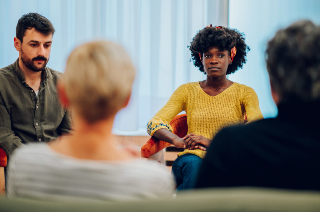 Diverse group of patients gathered in circle at psychologist office discussing self problems while listening others. Getting psychological support. Focus on an african american woman with afro hair.