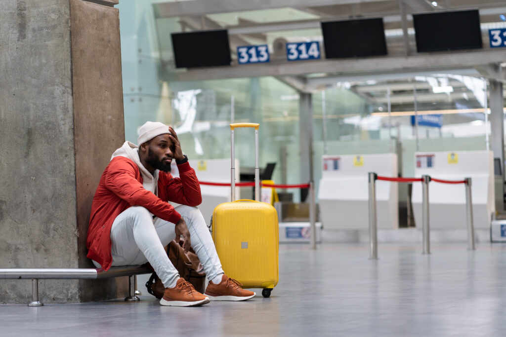 Sad African American man upset at airport his flight is delayed. Depressed traveler male waiting for a plane sitting in empty terminal with baggage. Exhausted guy on a long night connection at airport