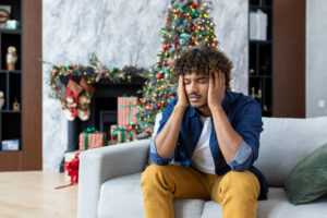 Severe hangover headache and migraines, man holding head sitting on sofa in living room for Christmas, African American man with curly hair near decorated Christmas tree.