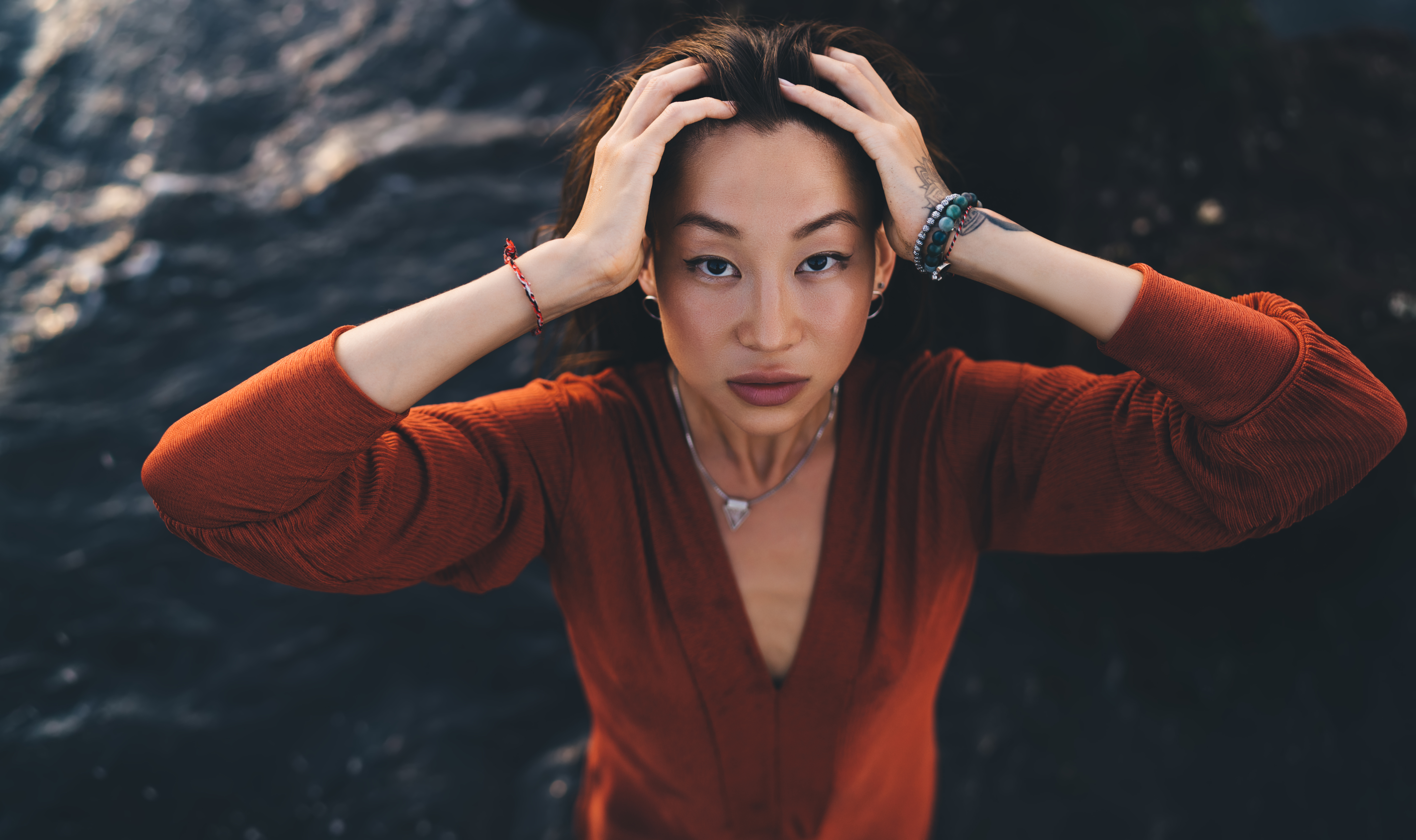 From above of young ethnic female model in stylish rust colored dress touching hair and looking up at camera against blurred dark blue ocean water and shore rocks in Bali
