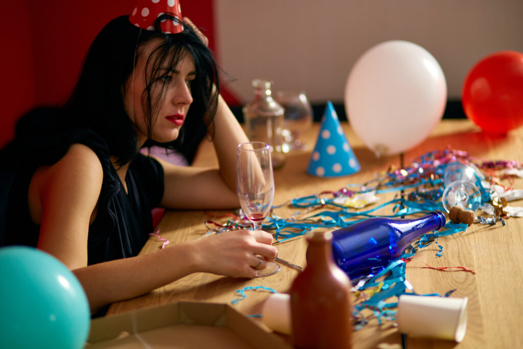 Young woman with red rubbed lipstick and cap, sitting tired at table in messy room after birthday party, woman after party at home