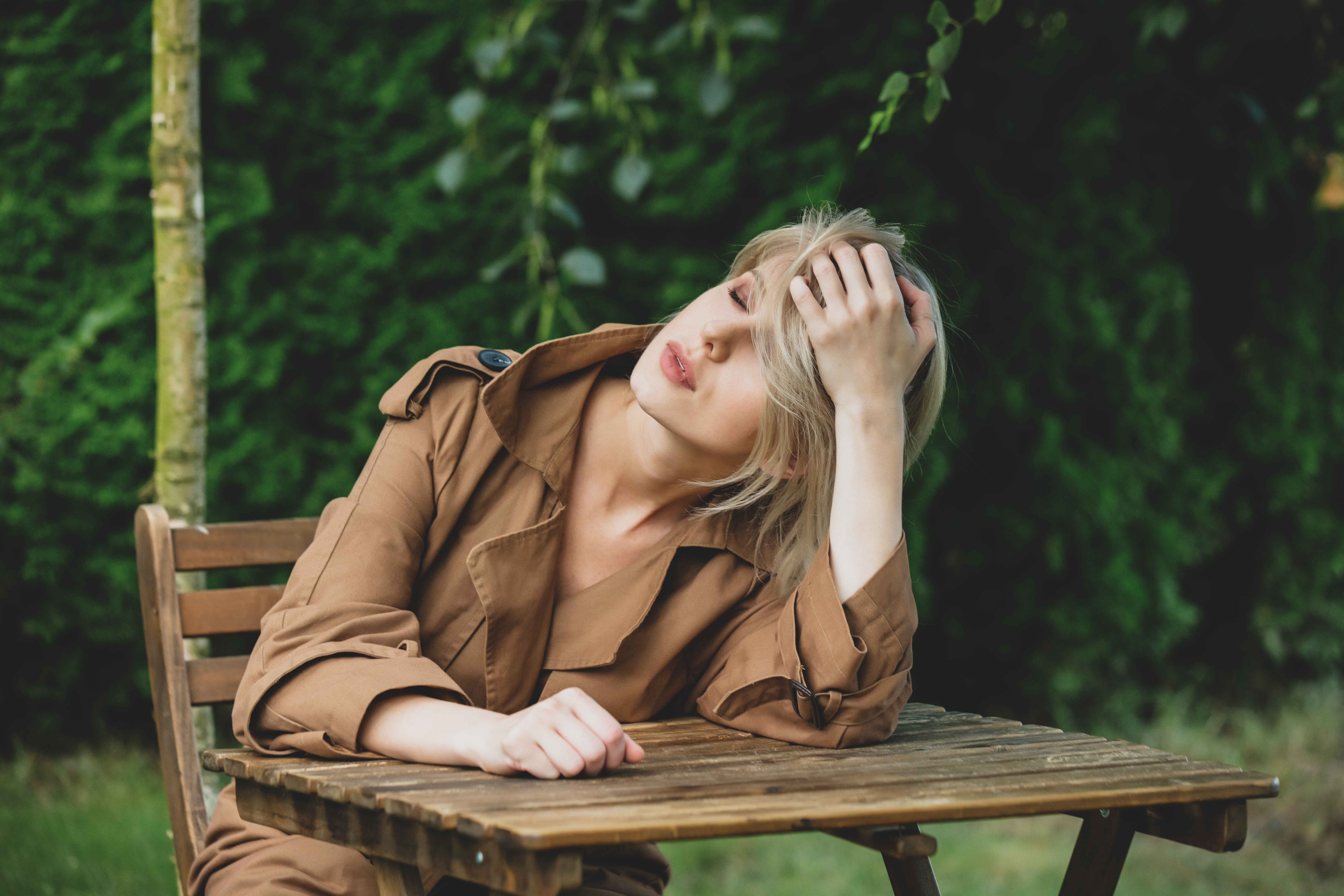 Beautiful woman in coat with blond hair sits at wooden table in a garden with trees on background