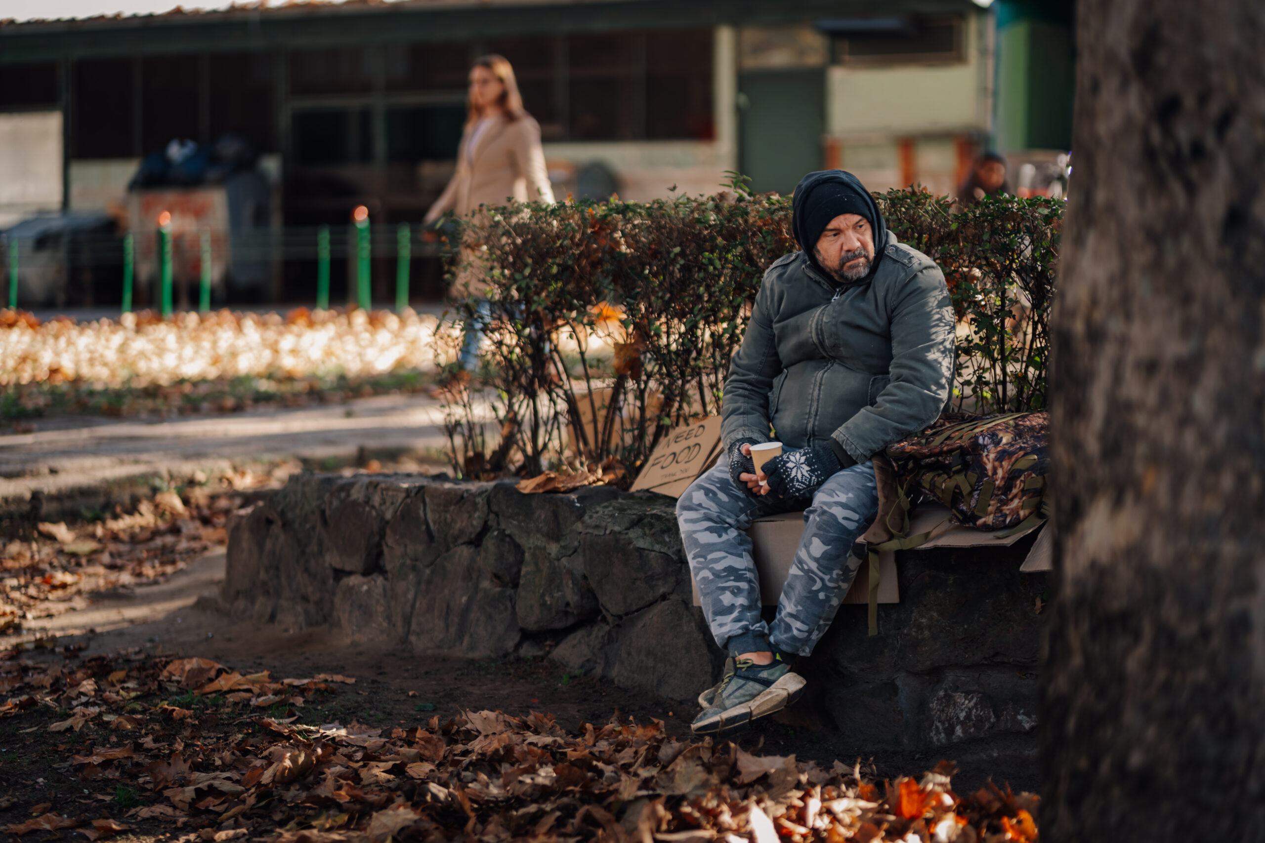 Homeless man is sitting on a stone wall, holding a cup, and displaying a cardboard sign asking for food in a park
