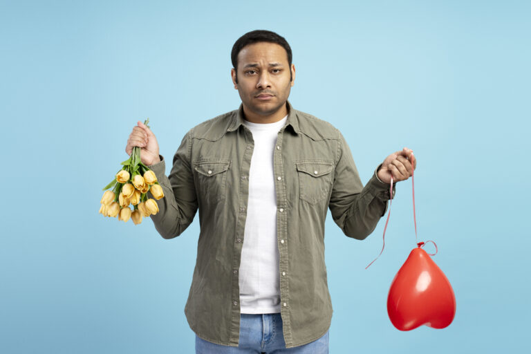 Disappointed man holding bouquet of yellow tulips and deflated red heart-shaped balloon, expressing sadness and failed romance on a vibrant blue background. Problem, divorce, disappointment concept
