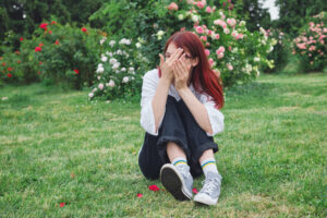 Teenage girl with red hair and glasses playfully hiding her face with hands while sitting on grass in a blooming garden